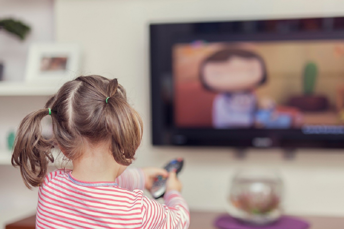 μικρό κοριτσάκι με κοτσίδες κρατά τηλεοκοντρόλ και βλέπει τηλεόραση